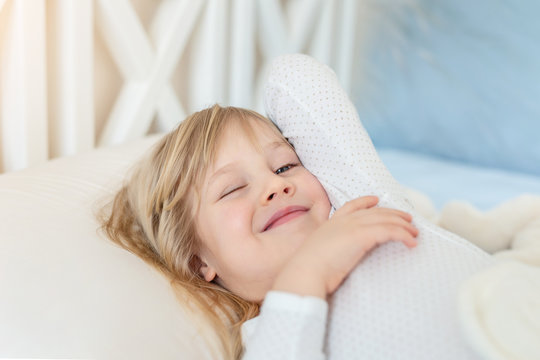 Cute adorable caucasian blond little toddler kid girl lying in bed on white pillow. Adorable sweet happy smiling child stretching wake up early morning for kindergarten or school. Childhood concept