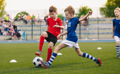Obraz premium Kids Football Players Kicking Ball on Soccer Field. Sports Soccer Horizontal Background. Spectators on Stadium in the Background. Youth Junior Athletes in Red and Blue Soccer Shirts. Sports Education