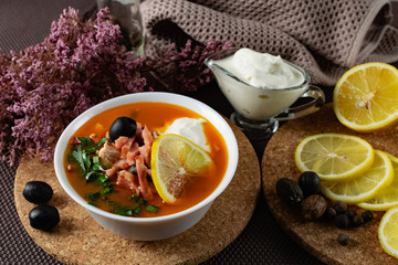 Soup Solyanka is a combined meat soup with smoked meat , various types of meat and tomato paste, serving dishes with sour cream, lemon and fresh herbs. Black olives on the table and a sprig of fresh f
