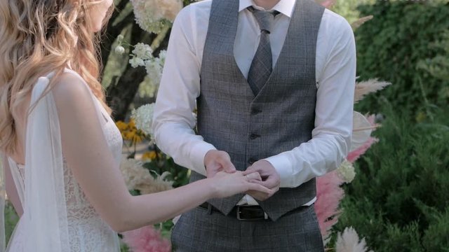 A White Groom At A Wedding Puts A Ring On His Bride Close-up Against An Arch Of Pampas Rose And Yellow Grass And Roses, On A Summer Day