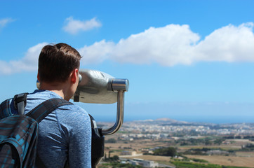 Fototapeta na wymiar Young tourist man in hat looking at Malta view through telescope. Horizontal shot. Copy space. 