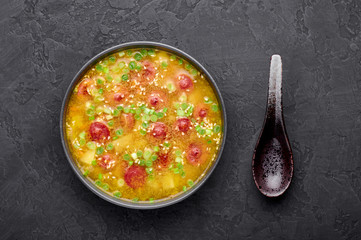 Dry Pea Soup with meat sausages, potato, green onions and sesame. Yellow Pea Soup in black bowl at dark slate background.