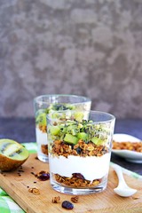 Homemade baked oat granola with nuts, dried cranberries and honey and unsweetened natural yogurt and kiwi in a glass glass on a dark concrete background. Healthy breakfast.