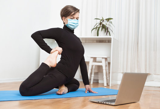 A woman in a medical mask does yoga through an online video conference on a laptop at home.