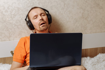 A young man in an orange T-shirt works at a laptop in headphones at home while lying on a bed. I fell asleep from fatigue. Remote work.