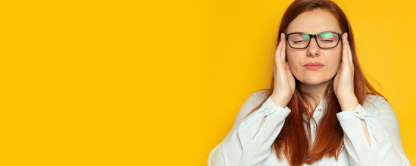 Young woman working woman with red long hair and glasses suffers a headache, holding his head on a yellow wall background. Stress, fatigue people concept.