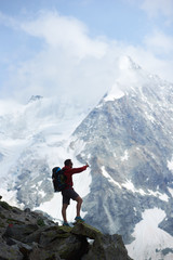 Male tourist with backpack looking at beautiful mountains scenery and pointing with finger. Man reaching peak, standing on stones. Wild nature with amazing views. Sport tourism in Alps.