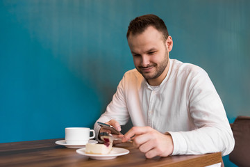 Young attractive businessman in a white shirt sits at a table in the phone and smiles, drinks coffee and eats cake on a blue background