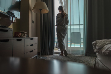 Silhouette of a young pretty woman which is standing in the white bathrobe and is looking on the sea view from her hotel room early at the morning