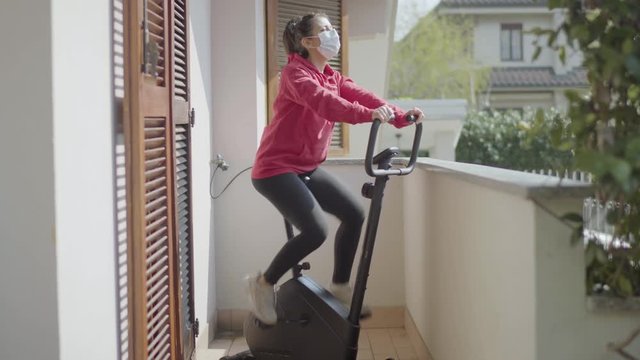 Woman with surgical mask pedaling exercise bike on the balcony