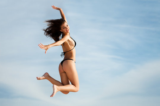 Happy girl in black swimsuit jumping high and smiling. Full length photo in motion with blue sky background