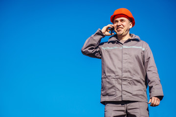 Construction worker in a helmet against the blue sky talking on the phone. Contractor control according to plan. Builder engineer helmet works at construction site. Copy space.