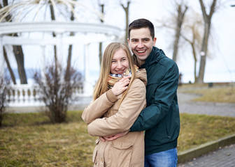 Young brunette man, wearing green jacket, standing behind young blond woman, wearing beige jacket, hugging her, smiling, kissing. Romantic Valentines day celebration