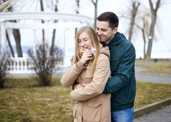 Fototapeta premium Young brunette man, wearing green jacket, standing behind young blond woman, wearing beige jacket, hugging her, smiling, kissing. Romantic Valentines day celebration