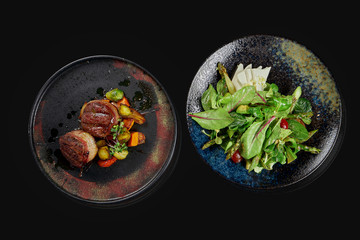 Two appetizing dishes - salad with feta cheese and tomatoes and beef steaks in stylish ceramic plates on a black background. Top view. Food photo for menu