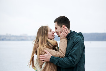 Young couple in love, wearing beige and green casual jackets, standing in front of frozen lake hugging, kissing each other in winter. Valentines day romantic celebration outside