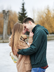 Fototapeta premium Young couple in love, wearing beige and green casual jackets, standing in front of frozen lake hugging, kissing each other in winter. Valentines day romantic celebration outside