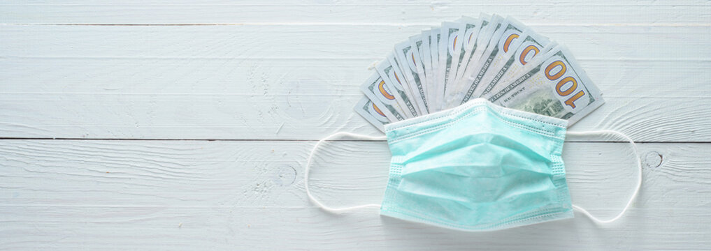 Dollars In A Medical Mask. Shot From Above On A White Wooden Background.