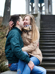 Young blond woman, is sitting on brunette man's lap, hugging each other and kissing on the old stairway. Romantic Valentines day celebration