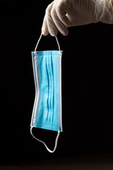 sanitary mask with one blue face and the other white with two elastic bands, held by one hand with a clear sanitary glove, on a black background