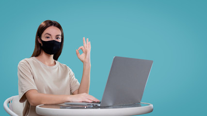 Pretty brunette female works with her computer isolated, she shows ok sign and wear medical mask, isolated over blue bg