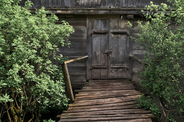 Old wooden grey hut in the village. Old time watermill among green trees in coutryside. Doors of ancient wooden building,