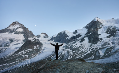 Happy traveller welcoming night in the mountains, standing with his back to the camera with open hands looking at glacier and high snowy peaks of Pennine Alps