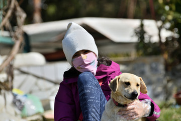 Girl with protective mask on her face sitting on the grass embraced with her dog