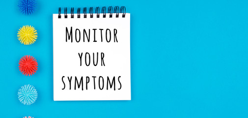 Paper notebook with plastic balls as viruses on the blue background with Monitor your symptoms wording. Epidemic, social isolation, coronavirus COVID-19 concept. Wide screen banner format