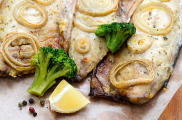 closeup fresh tender baked mackerel with rings of chopped onion, broccoli and lemon on a dark wooden cutting board in baking paper. keto and flexitarian diet and healthy eating.