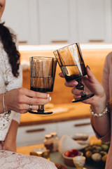Cropped stock photo of two unrecognizable friends celebrating event. Women clinking two glasses of wine in the kitchen.