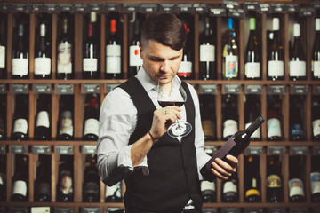 Bartender sniffs red wine in glass, holds a bottle, stands on background of cellar shelf.