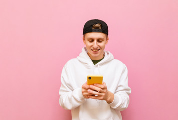 Portrait of cheerful young man in white hoodie and cap stands on pink background with smartphone in hand and uses internet. Guy teenager in casual clothes uses smartphone, isolated.