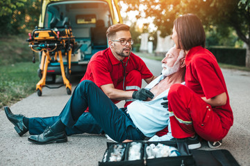 Elegant senior man with heart attack symptoms sitting on the road emergency medical service workers...