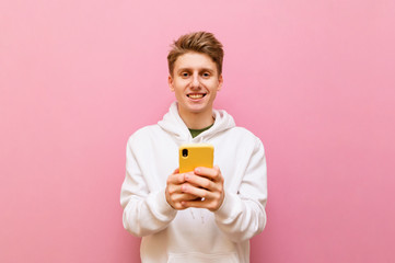 Cheerful blond guy in white casual clothes stands on a pink background with a smartphone in his hands, looks into the camera and smiles. Positive teenager using smartphone ,isolated. Copy space