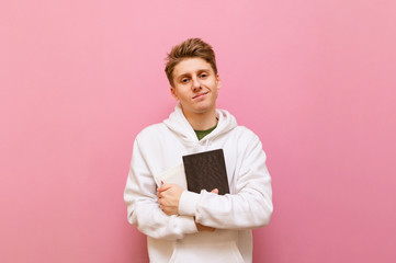 Portrait of handsome student boy with blond hair and in white hoodie stands on pink background and poses at camera with books in hands. Young blond man holding book in hand isolated on pink background