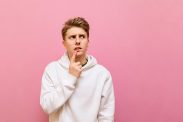 Thoughtful guy in white roe deer stands on a pink background and looks away at copy space with a serious face. Serious young man with blond hair looks away and thinks. Isolated.