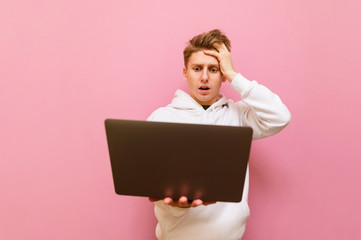 Shocked young man in casual clothes stands with laptop in hands on pink background and looks at computer screen with surprised face. Surprised student uses a laptop, isolated on a pink background