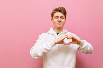 Positive handsome guy in a white sweater stands on a pink background and shows a gesture of love with a smile on his face, looking into the camera. Cheerful guy shows gesture of heart and laughs.