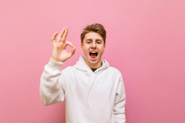 Fototapeta premium Happy guy in white casual clothes stands on a pink background, shows his thumbs up gesture OK, and looks in camera with a smile on his face.Smiling young man showing thumbs up gesture 
