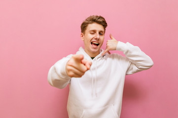Smiling young man in white sweatshirt shows call me gesture isolated on pink background. Positive...