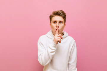 Concentrated young man stands on a pink background, looks into the camera and shows a gesture of silence. Handsome young man gestures shsh, finger on lips and looks intently at the camera, isolated.