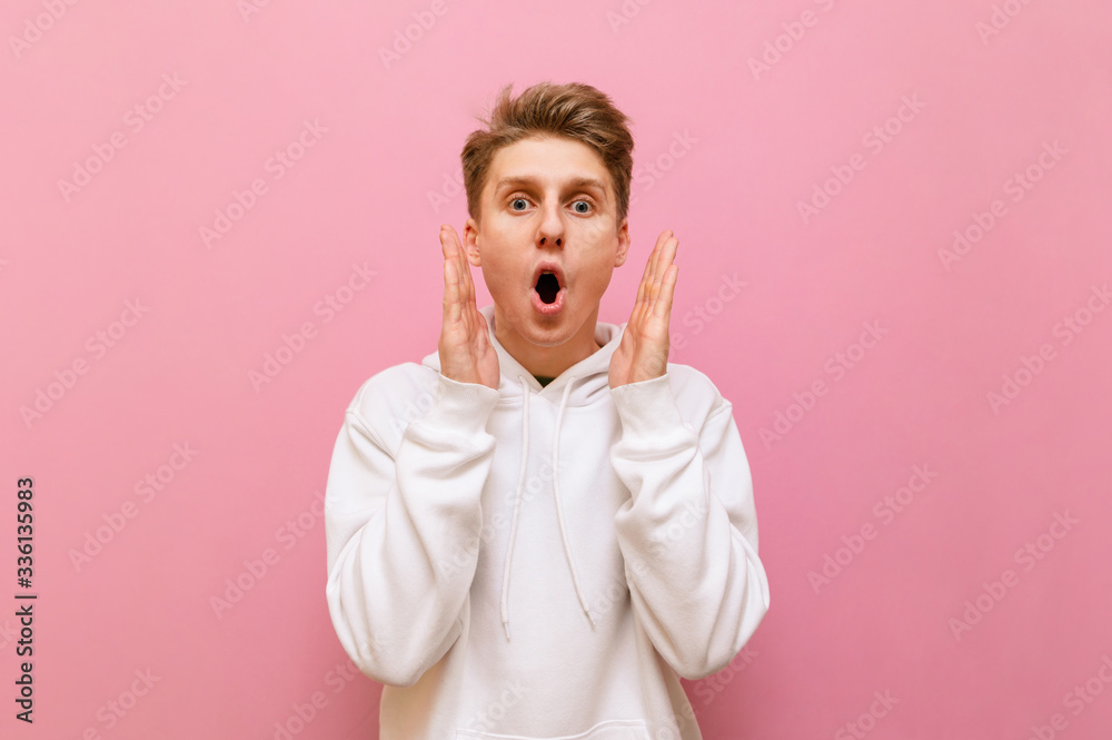 Wall mural Shocked young man standing on a pink background, looking mad at the camera and putting his hands to his face. Portrait of surprised guy in white hoodie looking into camera with crazy face. Copy space