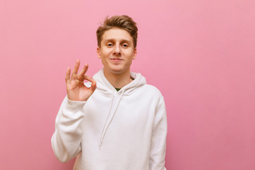 Cute young man in white casual clothes looks into the camera with a smile on his face and shows a...