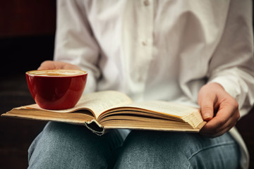 Woman with cup of coffee reading book indoors, closeup