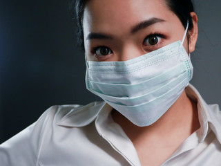 Head and shoulders portrait of Asian woman wearing protective mask and looking at camera posing against dark background