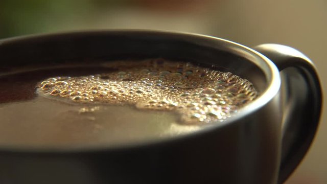 The dark coffee drink in a black mug with vapors, foam in golden color and bubbles. Steam above hot drink in macro view. 