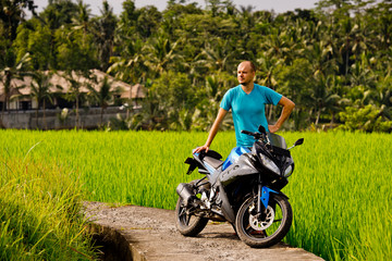 Plakat A young man standing near motorcycle and looks at him, in rice field.