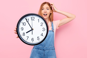 A shocked , nervous young lady on an isolated pink background holding a wall clock.