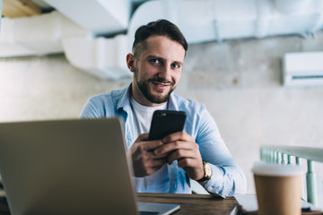 Obraz premium Portrait of prosperous male freelancer satisfied with earning online checking making account and transaction on smartphone, happy hipster guy student learning using modern devices in coffee shop